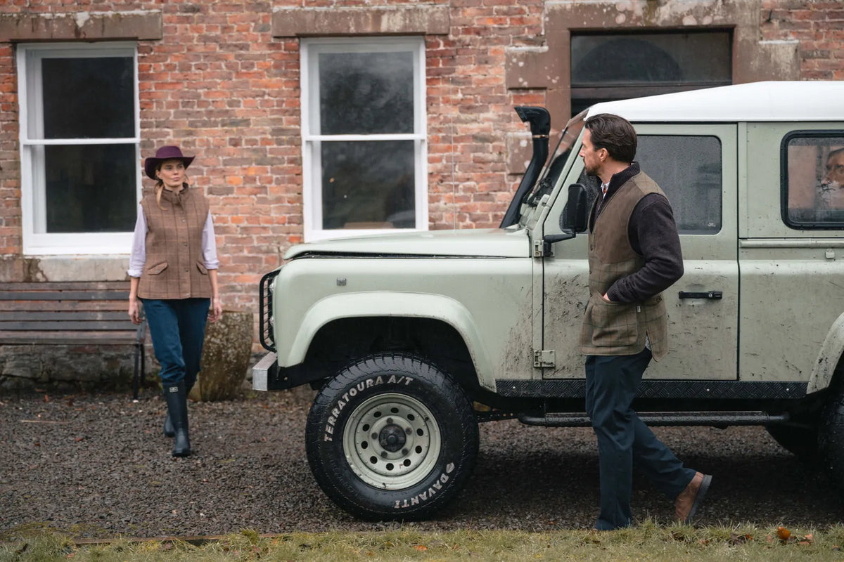 Two people wearing tweed gilets beside a Land Rover outside a country building in wet autumn conditions
