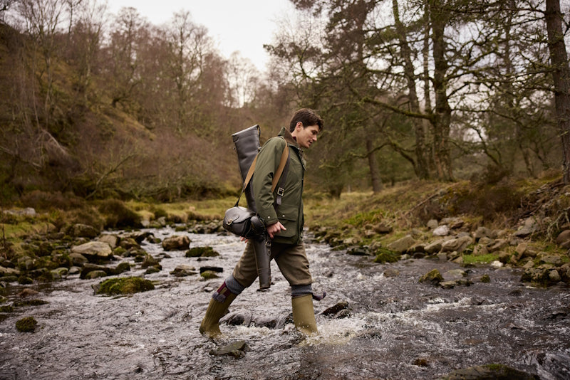 Shooter in a waterproof jacket and wellingtons crossing a river on country ground