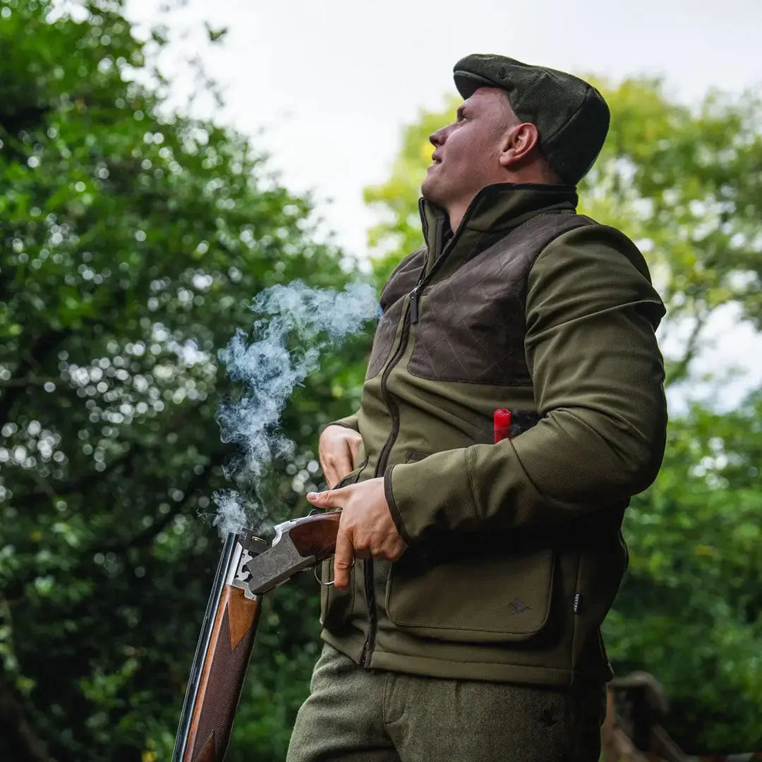 Shooter in a cap and shooting vest opening a break-action shotgun in a woodland setting